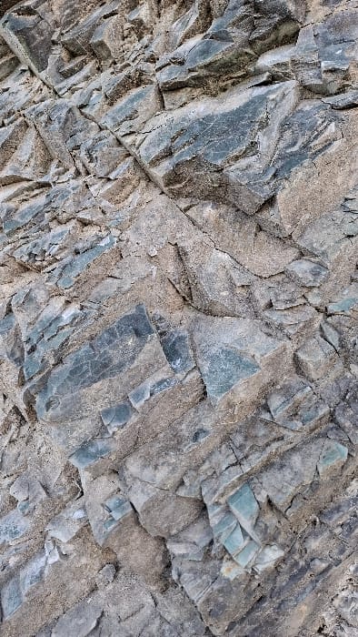 The image displays a detailed view of a fractured rock face in Podbadac, Montenegro. The composition is vertically oriented, revealing an intricate geological formation. The primary subjects are angular rock fragments and interstitial granular material. The rock face presents a dual coloration, featuring grey-brown weathered rock and distinct bluish-green mineral formations. The bluish-green components are often blocky or tabular, embedded within or partially covered by the lighter grey-brown matrix. Extensive cracking and fissures are evident across the entire surface, contributing to a rough, uneven texture indicative of natural stratification and erosion. No individuals, flora, fauna, or man-made objects are visible within the frame.