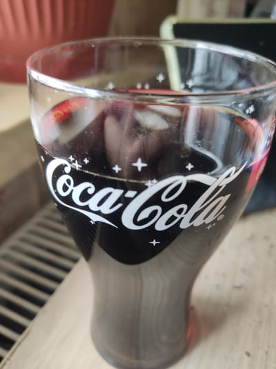 The image shows a Coca-Cola glass. The glass has a black background with the Coca-Cola logo in white. The background has some '+' symbols around the logo. The glass appears to be on a table. The location is Pavlovo, Russia.