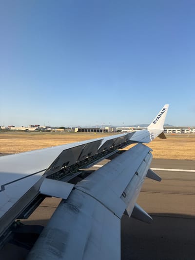 The image depicts a view from an airplane window, showing a portion of the aircraft's wing and its flaps extended on a tarmac. The wing is white with visible rivets and some smudges. The flaps are lowered, revealing mechanical components underneath. Beyond the wing, a runway is visible, with a yellow-brown, dry, grassy field bordering it. In the distance, airport buildings, hangars, and other aircraft are present under a clear blue sky. The airline logo "RYANAIR" is visible on the wingtip. The scene appears to be at Cagliari-Elmas Airport (SUJ) in Elmas, Italy, given the characteristic dry landscape and airport structures. There are no people visible in the image. The overall impression is a ground operation or preparation for takeoff/landing.