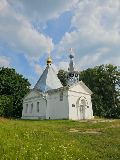 The image shows a white church with a gold cross on its roof, surrounded by lush green trees and a grassy field. The sky is blue with scattered white clouds. The church has a traditional architectural style, with a bell tower and a pointed roof. The scene appears to be set on a bright, sunny day. There is no visible text in the image. The image is taken in Ulitkino, Russia.