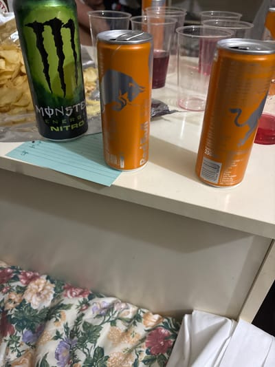 The image displays an indoor scene with various objects resting on a light-colored, possibly white or off-white, flat surface resembling a table or counter. The primary subjects are three energy drink cans: one green Monster Energy Nitro can on the left, partially obstructed by a translucent bag of potato chips, and two orange Red Bull cans positioned to its right. One Red Bull can is open, while the other appears to be closed. Several clear plastic cups, some containing residual dark red liquid, are scattered in the background. A crumpled piece of silver foil and a small, light blue rectangular piece of paper with faint writing are also visible on the surface. Beneath the table, a floral-patterned fabric, likely a bedspread or blanket, with prominent red, orange, purple, and green flowers on a cream background, is visible. The scene suggests the aftermath of a casual gathering or consumption of snacks and beverages. No individuals are clearly present. The image provides no specific visual cues or landmarks to identify a location within the city of Foligno, Italy.