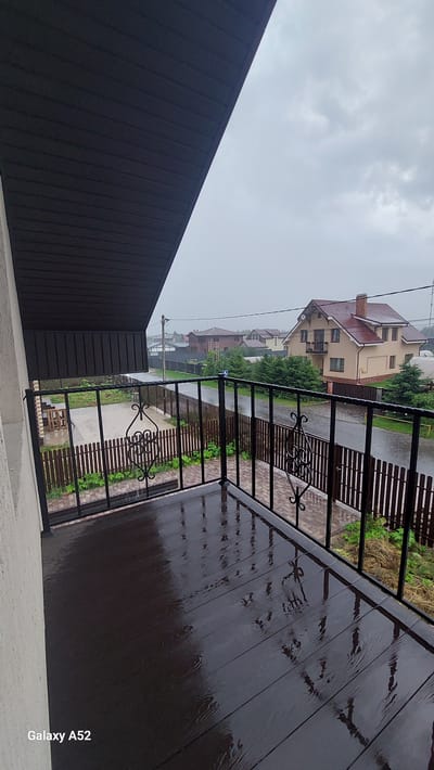 The image shows a wet, wooden balcony overlooking a street in a residential area during a rain shower. The balcony floor is dark brown and reflects the sky and surrounding objects due to the wetness. A black metal railing with ornate scrolling decorates the edge of the balcony. Beyond the railing, a wooden fence encloses a yard with some patchy grass and dry vegetation. A paved driveway is visible. Across the street, houses with various roof styles and colors are nestled among green trees. The sky is overcast and gray, indicating rainy weather. The scene appears to be located in the city of Dolche Vita KP, Russia, as suggested by the device watermark in the bottom left corner, "Galaxy A52". There are no people visible in the image.