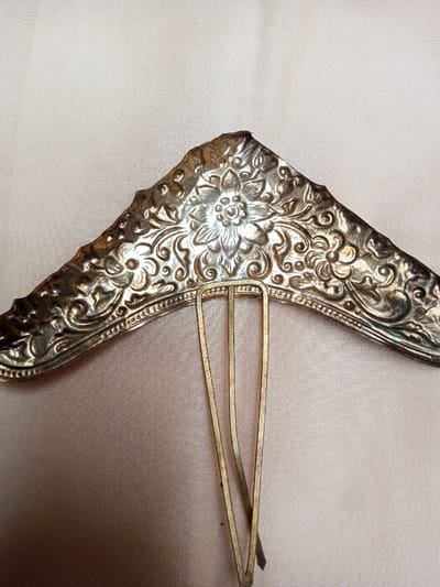 An ornate metallic hair comb is presented in a detailed close-up, presumably situated in Qarabucaq qəsəbə, Azerbaijan. The comb's decorative headpiece is constructed from a silver-toned metal, featuring a prominent central six-petaled floral design surrounded by intricate embossed scrollwork and additional smaller floral motifs. The upper edge of the headpiece is irregularly scalloped and undulating, forming a broad, curved "V" shape with a central apex. The metal surface exhibits areas of tarnish and minor surface wear consistent with age. Three slender, brass-toned metal prongs extend downwards from the underside of the decorative headpiece. These prongs appear slightly splayed at their tips. The entire object rests upon a light beige, subtly textured fabric background, which shows minor folds. No individuals, actions, or other objects are visible in the scene.