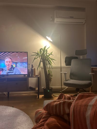 The image depicts an interior scene, likely a living room, in Karşıyaka, Turkey, illuminated by artificial light. A large flat-screen television on a light-colored media console dominates the left side, displaying a male individual in a grey jacket, possibly a reporter, against a background suggesting an indoor sports or entertainment facility. To the right of the television, on a small dark-framed side table, are three glass bottles, two clear and one amber. A tall, potted yucca-like plant stands between the side table and a white, horizontally-finned radiator mounted on the wall. Adjacent to the plant is a tall, minimalist floor lamp with a bright, upward-angled light source, casting a significant glow on the wall. Further right, a grey ergonomic office chair with a headrest is visible. Above the radiator and chair, a white Electrolux wall-mounted air conditioning unit is affixed to the wall, displaying a small red indicator light. A light-colored wooden floor covers the visible area, and a white or off-white wall forms the background. In the lower right foreground, an orange and white striped textured blanket partially covers an unseen object or person, suggesting a relaxed viewing posture.