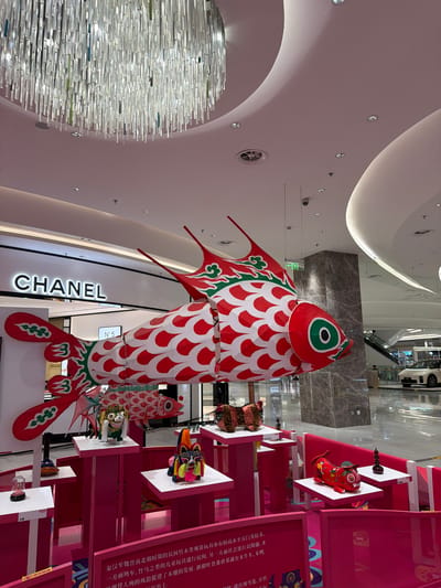 The image depicts a large, red and white decorative fish lantern suspended from the ceiling of a modern indoor shopping mall. The fish is elaborately decorated with red scales and green accents on its fins and head. Below the fish, a display of smaller, colorful traditional Chinese cultural figures, including what appear to be lion dancers and tiger figurines, are arranged on pink tiered platforms. In the background, a prominent "CHANEL" logo is visible on a storefront, suggesting a luxury retail environment. Further in the background, escalators and a white car are visible, indicating a spacious and contemporary mall interior. The overall scene suggests a festive or cultural display, possibly related to a holiday or celebration. The text visible on the pink displays is in Chinese, implying the location is in China. The presence of the "CHANEL" store and the modern architecture suggest a high-end shopping complex. The image appears to be taken from a slightly low angle, looking up towards the suspended lantern and the mall ceiling. The lighting is bright and diffused, typical of an indoor commercial space.