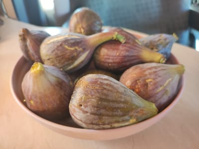 A shallow, light pink bowl is filled with ripe, dark purple and green figs. The figs are plump and vary slightly in color, with some showing hints of green and others deep purple. A few have visible stems. The bowl is placed on a light-colored, textured surface that appears to be a tablecloth or placemat. In the background, out of focus, are blurred shapes of furniture and possibly windows, suggesting an indoor setting. The lighting appears natural and soft, possibly indicating daytime. The scene is a simple still life focusing on the fresh fruit. There is no visible text. The location context of Valverde de Leganés, Spain, suggests this could be a home or a market.