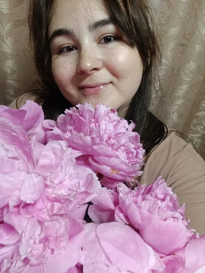 A woman with brown hair and freckles smiles at the camera, holding a bouquet of pink peonies in front of her. She is wearing a beige t-shirt. The background is a textured, beige curtain with a floral pattern. The lighting suggests it is daytime, possibly indoors. The woman appears happy and relaxed. There is no visible text in the image. The setting is likely a home or indoor space in Усть-Тунтор, Russia.