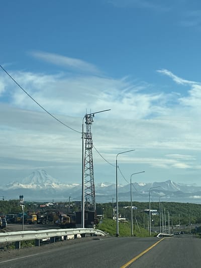 The image shows a view of a road leading towards a distant mountain range with snow-capped peaks under a partly cloudy blue sky. In the foreground, a paved road with a yellow dividing line curves to the right. A white guardrail runs along the left side of the road, with a grassy embankment behind it. To the left, a tall, lattice-style tower, likely for telecommunications, stands prominently. In the middle ground, there is a collection of heavy machinery, including yellow construction trucks and other vehicles, parked in an industrial or storage area. Beyond the machinery, several streetlights line the side of the road. Further in the distance, a treed hill rises, and beyond that, the snow-covered mountains are visible. The location is identified as Крутобереговый, Russia. There are no people or animals clearly visible in the image.