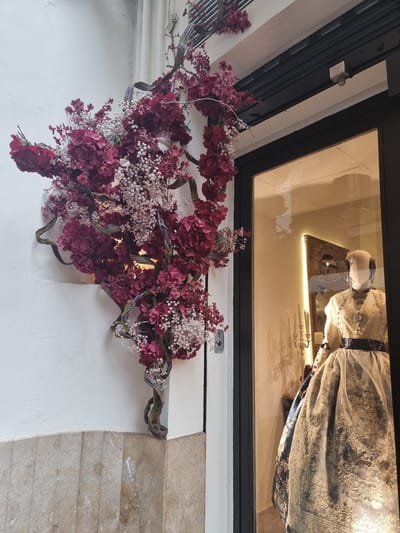 The image displays a detailed architectural facade featuring a display window and a decorative floral arrangement.

**Subjects:**
The upper portion of the image features a rectangular display case or window with a black frame. Inside, an effigy or mannequin is visible, lying down and dressed in an ornate, light-colored gown. The gown is rich with intricate lace or embroidered patterns and appears to be of historical or religious significance. The figure wears a head covering. Reflections in the glass show what appears to be a wooden structure or altar and other faint decorative elements, possibly candlesticks, illuminated by an internal light source.
Below the display case, a prominent and elaborate floral decoration is mounted on a wall. This arrangement consists of numerous clusters of dark red or burgundy flowers, resembling hydrangeas, interspersed with delicate sprays of tiny white flowers, likely baby's breath. These flowers are woven into gnarled, dark branches, creating a natural, flowing design.

**Setting and Details:**
The display case and floral arrangement are set against a white, textured wall. To the right, a section of the wall is covered with horizontal, light-brown stone or tiled blocks. Above the display, portions of a dark overhead structure, possibly an awning or shutter, are visible, along with a small, light-colored metal fixture attached to the ceiling.
The overall scene suggests a public display, possibly belonging to a historical or religious institution, or a shop specializing in traditional goods, consistent with the cultural context of Xàtiva, Spain. There is no explicit activity taking place, and no visible text. The lighting suggests an indoor or shaded outdoor environment, with the display case specifically illuminated.