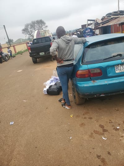 The image depicts an outdoor scene on a cloudy day, likely in Ohit, Nigeria. In the foreground, a person wearing a grey hoodie and blue jeans leans against the back of a teal-colored hatchback car. The car has visible wear and tear, including chipped paint on the rear bumper and a license plate with the characters "BC13". A pile of white clothing and a black plastic bag are on the ground next to the car. In the background, a black pickup truck is parked, with its license plate reading "NA 9548DD". Further back, there are various structures, including a building with a Goldberg beer advertisement and a row of shops with corrugated metal roofs. Other people are visible in the background, and there is a general sense of activity in a semi-urban or roadside environment. The ground is paved with a reddish-brown, gravelly material, and there is scattered litter.