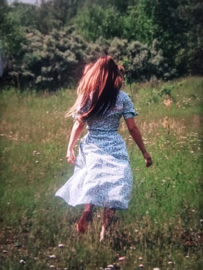 A young woman with long, auburn hair is captured from behind, running through a grassy field. She is wearing a light blue, floral-patterned dress with puff sleeves. The dress is flowing around her as she runs. Her feet are bare and visible as they move through the grass. The environment is a natural, outdoor setting with a backdrop of dense green trees. The field is dotted with small white wildflowers. The lighting suggests it is daytime, likely a sunny or partly cloudy day. The image is taken from a medium distance, focusing on the woman as she moves away from the viewer. The image does not provide any specific indicators to determine its location in the city of Serov, Russia.