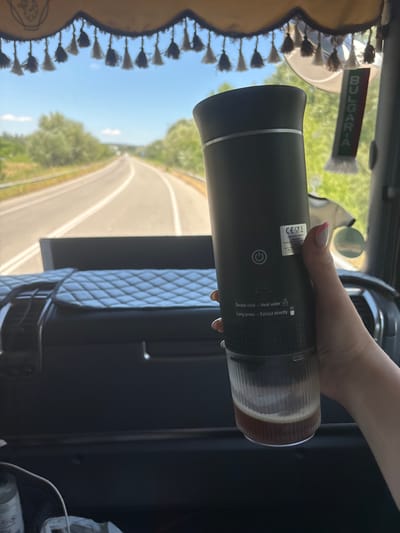 A woman's hand holds a black portable coffee maker containing a dark liquid with a foamy top. The coffee maker is held in front of a windshield with a view of a road lined with trees and a bright blue sky. A fringed valance hangs from the top of the windshield, and a small Bulgarian flag keychain dangles from the rearview mirror. The interior of what appears to be a truck is visible, with a quilted dashboard cover and a steering wheel. The scene is set on a sunny day, likely during travel.