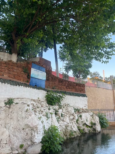 The image depicts the Ain Khadem spring in El Hajeb, Morocco, a natural water source with historical and cultural significance. A blue sign with Arabic script is affixed to a red stone wall, indicating the spring's name. The spring emerges from a rocky embankment covered in white plaster and greenery, with water flowing into a pool in the foreground. Overhanging trees with lush green foliage provide shade. A white fence separates the spring area from a beige brick wall in the background, where a cellular tower is visible in the distance. The scene suggests a tranquil, natural setting within or on the outskirts of the city.
