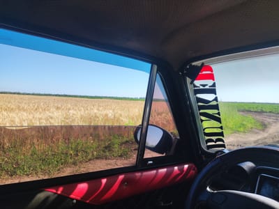 The image depicts the interior of a vehicle, looking out the side window towards a rural landscape. A field of ripe wheat stretches across the horizon under a clear blue sky. To the right, a dirt road curves through a green field. The interior of the car features a red upholstered door panel and a portion of the dashboard, including the steering wheel and an instrument cluster. A black sticker with white lettering, partially obscured by the window frame, is visible on the right side of the windshield. The scene is illuminated by bright sunlight, casting shadows inside the car. The specific location in Mykolaivske, Ukraine, is not directly indicated by any visible landmarks or signs.