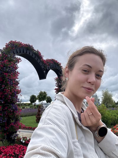 A young woman with blonde hair pulled back is taking a selfie outdoors in a garden. She is wearing a white corduroy jacket. Her left hand is raised to her face, forming a finger heart gesture. She is wearing a ring on her left ring finger and a smartwatch on her left wrist. Behind her, a large heart-shaped arch is covered in pink and purple flowers. The background shows a park with trees, manicured bushes, and a cloudy sky.