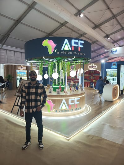 The image shows a man standing in front of a booth at what appears to be a trade show or exhibition. The man is wearing a plaid shirt, dark jeans, and sneakers. He has a beard and is wearing a baseball cap.
Behind him is a large, circular booth with the letters "AFF" displayed prominently. The booth also has a logo resembling the African continent in shades of green, pink, and white. The phrase "a vision to share" is beneath the letters. The booth has hanging plants and glowing orbs. It appears to be illuminated with LED lighting.
In the background, other vendor booths can be seen, including one with the name "Kayna" and another with the name "Jibal." People are visible in the background, some engaged in conversation.
The location is likely the city of Al Machouar – Stinia, Morocco, as indicated in the prompt, although this is not visually verifiable from the image alone. The environment suggests an indoor venue, possibly a convention center or exhibition hall.
