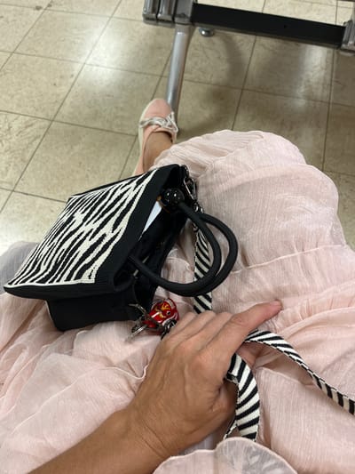 The image shows a close-up view of a person's lap and handbag, with the background out of focus. The person is wearing a light pink, sheer dress and pink slip-on shoes. A black and white zebra-patterned handbag with a round black handle and a detachable striped strap is resting on the person's lap. A small red and yellow charm, possibly a Daruma doll, is attached to the handbag. The person's left hand is visible, holding the handbag strap. The scene appears to be indoors, on a tiled floor, possibly in a waiting area or public space. The overall impression is one of casual waiting. There is no indication of the specific location within Katunayake, Sri Lanka.