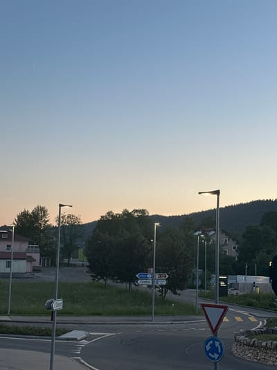 This is a vertical, eye-level, medium shot of a street in Le Brassus, Switzerland, at dusk. The sky transitions from a pale blue to a soft orange, suggesting the sun has recently set or is about to rise. Streetlights are illuminated, casting a warm glow on the scene. A winding road curves to the right, marked with yellow and white lines.

On the side of the road, several signs are visible. One is a triangular "give way" sign with a blue circular sign showing three arrows indicating a roundabout. Another sign displays directional arrows with text that is partially legible but appears to include "Centre" and possibly "La Russes." Further down, a street sign reads "L'Orient."

Behind the road and signs, there is a grassy area bordered by trees. In the background, a hillside is covered with dense trees, and a building with a light-colored facade and dark windows is nestled amongst them. Another building, with a lighter, modern design, is visible closer to the foreground on the left. The overall atmosphere is calm and quiet, characteristic of a twilight in a semi-rural setting.