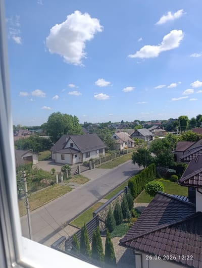 This is a high-angle, medium-shot photograph taken on a sunny day with scattered white clouds against a bright blue sky. The scene depicts a suburban residential area, likely in Novovolynsk, Ukraine. In the foreground, the edge of a white window frame partially obstructs the view.
The middle ground features a paved road winding through a neighborhood of detached houses. Many of the houses have dark brown tiled roofs and are surrounded by green lawns, trees, and fences. Some houses have visible windows and architectural details like chimneys. A tall concrete utility pole with wires runs along the side of the road.
The background shows more houses and dense green trees, creating a sense of a well-established, leafy community. The overall impression is one of a peaceful, ordinary day in a residential area during daylight hours. The weather appears clear and warm.
Visible text in the bottom right corner reads "26.06.2026 12:22", indicating the date and time the photograph was taken. There are no people visible in the image.