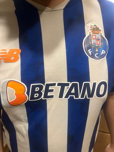 The image displays a torso-level, front-facing view of an individual wearing a blue and white vertically striped FC Porto home jersey. The orange "New Balance" (NB) logo is present on the upper left chest. On the upper right chest, the circular FC Porto club crest is visible. Centered on the jersey is the "BETANO" sponsor logo, rendered in blue lettering with a white outline, accompanied by an orange and white "B" emblem to its left. The jersey fabric exhibits a subtle textured pattern. A portion of the individual's neck is visible at the top, and their right arm is partially visible on the right side of the frame. The background is indistinct, light-toned, and out of focus, consistent with an indoor setting. This scene is located in Pedrouços, Portugal.