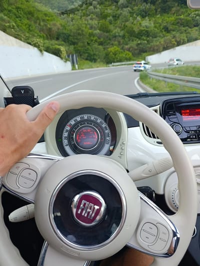The image shows a first-person perspective of someone driving a white Fiat 500 through a mountainous, tree-covered area. The driver's hand is on the beige steering wheel, which features the Fiat logo and various control buttons. The dashboard displays a digital readout showing the date as "29 Jun", the time as "19:38", and the car's mileage as "90980". The ambient temperature is indicated as 32°C, and the "ECO" mode is active. The radio display is on, showing "FM-BO PLAY Radio" and a website address.

Outside the car, a white concrete wall lines the road, which curves to the right. Further ahead, a police car and another vehicle are visible on the road. The surrounding hills are densely covered with green trees, suggesting a warm, possibly summer day. The time of day, 19:38, indicates it is late evening, and the lighting suggests it might be dusk or overcast. The scene appears to be a road in a rural or scenic area, possibly a highway or a mountain pass. The overall impression is one of a road trip in pleasant but potentially warm weather, with the driver engaged in navigating the route.