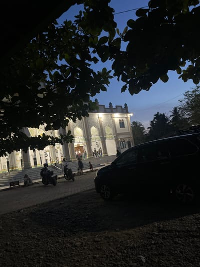 The image depicts a mosque in Lapang Timu, Indonesia, during twilight. The mosque's facade is illuminated, showcasing intricate white latticework and decorative panels against a beige background. A wide set of stairs leads up to the entrance, with several people gathered at the top and on the steps. In the foreground, a dark-colored SUV is parked on a gravelly surface. To the left of the mosque, a person in a hijab sits on the ground near a motorcycle. Other motorcycles are also visible, and a few individuals are standing or walking near the mosque's entrance. Lush green trees with dark foliage frame the top and left sides of the image, silhouetted against the darkening blue sky. The overall scene suggests a peaceful evening gathering outside a place of worship.