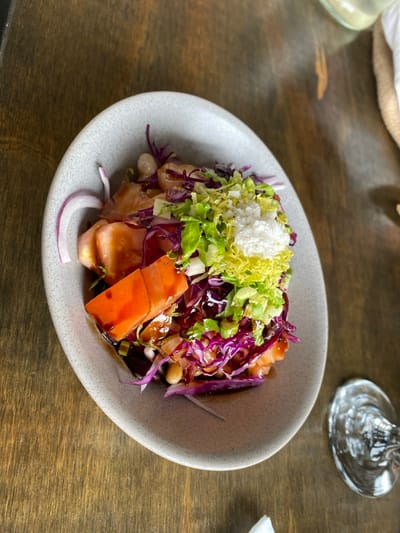 The image displays a meal served in an irregularly shaped, light grey speckled ceramic bowl, positioned slightly left of center on a dark brown wooden table with visible grain. The dish consists of several layered ingredients: light orange-hued protein (likely fish), chunks of reddish-orange vegetable, scattered white beans, shredded purple cabbage, finely chopped green lettuce, and a prominent mound of white granular topping (possibly grated cheese or salt) on top of the green lettuce. A crescent slice of red onion is placed at the left edge of the bowl. A dark, glossy liquid sauce or dressing is visible at the bottom of the bowl and coats some ingredients. To the lower right of the plate, a clear stemmed glass is partially visible. A small piece of white material, possibly a napkin, is at the bottom edge of the frame. The scene is set in a dining environment, likely indoors, within the city of Puerto Almanza, Argentina. There are no people visible.