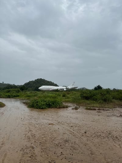 A large, white airplane is parked in a grassy, overgrown field. The sky is overcast and the ground in the foreground is muddy and wet, suggesting recent rain. The airplane is a Boeing 747, a wide-body commercial jetliner. It is positioned at an angle, facing towards the right of the frame. Behind the airplane, a green, tree-covered hill rises. The environment appears rural and somewhat neglected. The scene is devoid of people. The location is identified as Pak Nam Pran, Thailand.