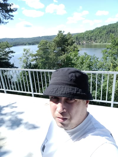The image is a selfie taken by a man wearing a black bucket hat and a white t-shirt. He is standing on a white surface, possibly a balcony or deck, with a white railing in the foreground. Behind him is a scenic view of a lake surrounded by lush green trees and hills. The sky is blue with scattered white clouds. The man's face is partially visible, and he appears to be looking at the camera. The overall impression is of a sunny day in a natural setting.
The location is Máchovo jezero (Lake Mácha) in Doksy, Czechia. The view suggests the selfie was taken from a vantage point overlooking the lake, such as a restaurant, hotel, or viewpoint on the lake's shore. The characteristic shape of the lake and the surrounding hills are identifiable features of this popular tourist destination.