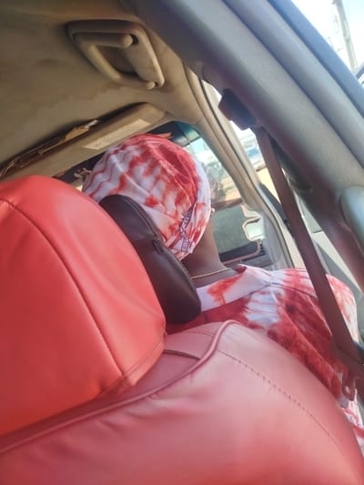 The image shows the interior of a car, with a focus on the back seat. A person wearing a red and white tie-dye headscarf and matching outfit is seated in the back. Their head is turned away from the camera, and a portion of their neck and shoulder is visible. They are wearing a gold chain necklace. The seat appears to be upholstered in red leather. A dark headrest is visible behind the person. The car's interior ceiling is a light beige color, and a handle is attached to it. A sun visor is also visible on the ceiling. The scene is brightly lit, suggesting it is daytime. The weather appears to be sunny. The background, visible through the car window, shows a street scene, with some vehicles and buildings. The image is taken from a low angle, looking up towards the ceiling of the car. There is some text visible on the sun visor, but it is too blurry to read. The location is specified as Okha, Nigeria.