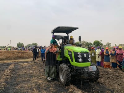 The image shows a green tractor with a "WOFAN" logo, surrounded by a group of people. The tractor is parked in a field of brown, dry grass. There is a large pile of stacked sticks in the background. A woman wearing a hijab and a patterned robe stands beside the tractor and is speaking into a microphone. A man in a white hard hat is visible at the driver's seat of the tractor. Other people are standing around the tractor, some holding bags or wearing hijabs. The scene appears to be a community gathering in an open field. The location is Ringimawa, Nigeria, with coordinates: 11.666 latitude, 8.415 longitude.