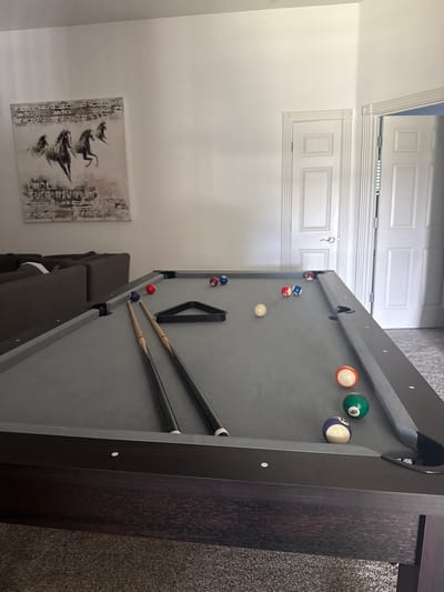 The image shows a billiard table in a room. The table is grey with dark brown wood trim. Several billiard balls are scattered on the table, along with two pool cues and a black ball rack. The room has light grey walls with a painting of horses on one wall. A brown couch is partially visible near the billiard table. The floor is covered in dark brown carpeting. A white door with raised panels is visible in the background. The location is in a room of a house in Southlake, United States of America.