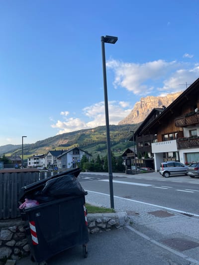The image depicts a street scene in La Ila - Stern - La Villa, Italy. In the foreground, a large black dumpster overflows with trash bags. To the right of the dumpster, a paved road with white lane markings stretches into the distance. Several cars are parked along the side of the road. Behind the road, a cluster of multi-story buildings with wooden balconies and potted plants line the street. In the background, a lush, green mountain slopes upwards, capped by a rugged, rocky peak illuminated by the sun. The sky above is a clear blue with scattered white clouds. A tall, modern lamppost stands in the middle of the scene, casting a shadow on the road. The overall impression is that of a quiet, picturesque mountain town.