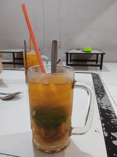 The image shows two clear glass mugs filled with a beverage, likely iced tea or a similar drink, on a white table. A spoon and an orange plastic straw are visible in the foreground mug. The mug in the foreground contains a green ingredient at the bottom. A second similar mug is in the background, also with a straw and spoon. In the background are two small black tables with a white top. The image appears to be taken indoors, with a white wall behind the tables. There is an electrical outlet on the wall behind the tables, and a green object on the table further away. The table is white with black and white decoration on the edge. Given the location, it's plausible this is in Cangkring, Indonesia. No people are visible in the image.