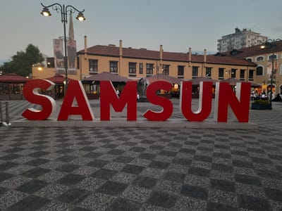 In Samsun, Turkey, the word "SAMSUN" is displayed in large red 3D letters on a plaza. The letters are positioned in front of a row of buildings, some of which appear to be restaurants with outdoor seating under umbrellas. In the background, a taller, modern building with multiple windows rises above the lower structures. Several people are visible in the plaza, some seated at tables and others standing. A lamppost with two lights is situated behind the "S" and "A" of the large letters, and banners featuring images of two individuals hang from the lamppost. The ground is paved with a dark, textured material arranged in a checkerboard pattern. The sky is overcast, suggesting it is either early morning or late evening.