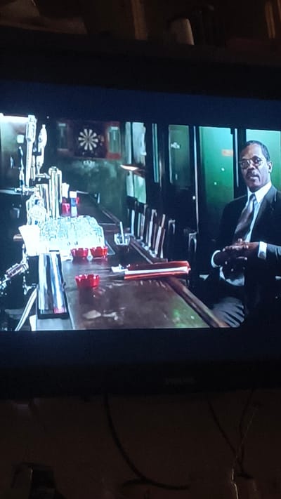 The image shows a man sitting at a bar. He is wearing a suit and tie and glasses. He is sitting with his hands clasped in front of him. The bar is made of dark wood and there are several bottles of alcohol behind the man. There is also a dartboard on the wall behind the bar. The scene appears to be taking place in a bar in Havdrup, Denmark, based on the inclusion of this location in the prompt.