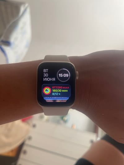 The image shows a close-up of a person's wrist wearing a light-colored smartwatch. The watch face displays the time as 15:09, and the date is June 30th, indicated by "30 ИЮНЯ" (30th of June). There are also activity tracking metrics visible, showing "677/260 ккал" (677/260 kcal), "180/30 мин" (180/30 min), and "8/12 ч" (8/12 h), suggesting progress towards goals for calories burned, exercise minutes, and standing hours. A circular progress indicator with red, yellow, and green segments is also present on the watch face, typical of fitness trackers. The background is blurred but appears to be an indoor setting with soft lighting, possibly a home environment. The overall impression is a snapshot of daily life, focusing on personal health and fitness tracking. The location context is Holíč, Slovakia.