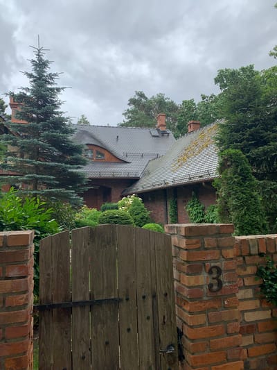 The image captures a view of a property entrance in Pobierowo, Poland. A rustic wooden gate, attached to a brick pillar on the left and a brick pillar with the number "3" on the right, stands closed. The gate is constructed from vertical wooden planks and features a dark metal hinge. Beyond the gate, a brick wall forms the boundary of the property. The main structure visible is a large, multi-gabled house with a dark tiled roof that shows some signs of moss growth. Red brick is a prominent material for the house's exterior walls, and there are visible chimneys on the roof. Lush green foliage, including mature evergreen trees, shrubs, and flowering bushes, surrounds the house and frames the entrance. The sky is overcast with a muted, cloudy appearance. The overall scene depicts a well-established residential property with a naturalistic garden setting.