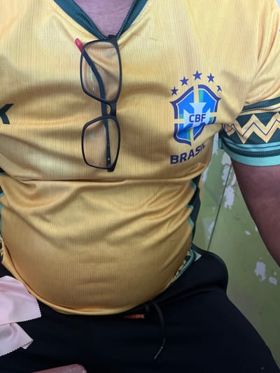 A person is wearing a yellow Brazil national football team jersey and black sweatpants. A pair of black-rimmed glasses is resting on the collar of the jersey. The person's belly is visible. The background consists of a light green wall with white splatters. The scene is indoors and appears to be a casual setting. There is no indication of the location being Langnibong, Indonesia.