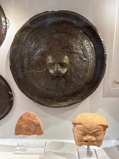 The image depicts an archaeological display within a museum, likely the Heraklion Archaeological Museum in Heraklion, Greece, given the nature of the artifacts.

The central and largest object is a circular, dark metallic shield, possibly bronze, with a prominent, raised, grotesque face at its center. The face has wide eyes, a flattened nose, and an open mouth. Surrounding the central face, the shield is intricately decorated with embossed patterns and what appear to be serpentine or draconic figures. The rim of the shield is rough and shows signs of age and wear.

To the lower left of the shield, a small, reddish-brown clay fragment is displayed on a clear acrylic stand. This fragment appears to be part of a larger object, possibly a vessel or sculpture, with faint markings suggesting a face or design.

To the lower right of the shield, a larger, light brown ceramic mask is mounted on a metal rod within a clear acrylic stand. This mask has a stern, somewhat exaggerated facial expression with deep-set eyes, a broad nose, and prominent cheekbones. The mask is detailed with lines suggesting hair or a headdress and some scarring or texture on its surface.

The objects are set against a plain, light-colored wall, indicative of a museum exhibit. The lighting appears to be artificial, illuminating the artifacts clearly. The overall scene conveys a sense of historical significance and the preservation of ancient artifacts.