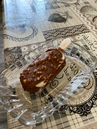 An ice cream bar with chocolate coating and nuts sits on a clear glass plate. The plate is on a patterned tablecloth. The tablecloth has a design of flowers and ornate borders, with a plaid pattern in some sections. The ice cream bar has a wooden stick protruding from the top. The background shows more of the tablecloth with various decorative elements. The scene is likely indoors, on a table. The image does not contain enough information to determine its location in Lebedinovka, Kyrgyzstan.