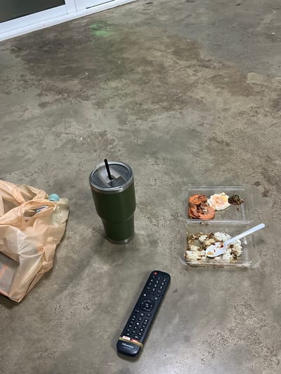 The image depicts an outdoor scene on a concrete surface, possibly in Thung Tako, Thailand, characterized by a weathered and stained ground. In the mid-ground, a green insulated tumbler with a black straw sits next to a light-colored plastic shopping bag, which appears somewhat rumpled and contains items within. To the right of the tumbler are two clear plastic food containers. The upper container holds three cooked shrimp and what appears to be a fried egg with some herbs. The lower container contains rice mixed with other ingredients, with a white plastic spoon resting on top. In the foreground, a black Hisense brand remote control, secured with two yellow rubber bands, is positioned diagonally. The background shows a white-topped structure, likely a doorway or window frame, and the concrete floor shows varying patches of darkness and lighter tones, suggesting moisture or stains, with small flecks of pink and green scattered across the surface. There are no people visible in the image.