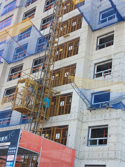 This is a medium-shot, eye-level, exterior, low-angle photograph of a construction site in Uiwang-si, South Korea. The image features a large, concrete building under construction, with numerous windows in various stages of completion. A yellow construction elevator is visible on the left side of the building, with a blue safety net covering the top of the structure. Orange safety netting and yellow scaffolding are also present. The building appears to be a multi-story residential or commercial complex, with numbered frames, indicating apartments or units, on the exterior. Signs with Korean text are visible on the construction elevator and on an orange barrier in the foreground. The overall scene depicts a typical urban construction environment.