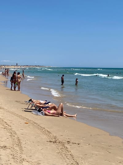 This is a landscape photograph of the beach in Conil de la Frontera, Spain. The image captures a sunny day at the beach, with a clear blue sky and gentle waves rolling onto the shore.
In the foreground, two women are sunbathing on lounge chairs on the sandy beach. One woman, wearing a pink bikini, is lying on her stomach on a beach towel. The other woman, wearing a white top and bikini bottoms, is lying on her back on a lounge chair.
In the middle ground, several people are walking along the shoreline and wading in the shallow water. A group of four women, some with children, are standing on the wet sand near the water's edge. Further out in the sea, several individuals are swimming and playing in the waves.
The background of the image shows a long stretch of coastline with a distant town visible on the horizon. The water is a clear turquoise color, and the waves are small and gentle. The sand on the beach has tire tracks from vehicles.
Overall, the image depicts a vibrant and active beach scene, typical of a summer day in Conil de la Frontera.
