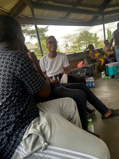 The image depicts an indoor-outdoor scene under a dark, corrugated metal roof supported by pillars, in Ozuaha, Nigeria. Multiple individuals are present, seated on a concrete bench and floor.
In the foreground, the back of a person wearing a black and white geometric patterned short-sleeved shirt is visible, facing left. Their legs, clad in light grey trousers with two white stripes near the cuff, are extended towards the bottom right. An Apple Fruit Milk bottle rests on the floor near their feet.
To their right, slightly further back, a young man with dark skin, black-framed glasses, and a short haircut is seated. He wears a white short-sleeved t-shirt, black trousers, and black slip-on sandals. He is smiling broadly, looking towards the person in the foreground. A white mesh wrap or bandage is visible on his left hand. A black bag with white accents is positioned across his lap, and he holds a dark object in his hands.
In the midground to the right, another person with dark skin is seated on a light-colored concrete bench, leaning back casually with their right arm resting on the bench's backrest. They are dressed in a vibrant yellow, blue, and red patterned traditional African outfit. A black shoulder bag rests next to them on the bench, along with an orange and white box.
Further right, partially visible, another individual wears a brown top or vest and blue jeans. On the concrete floor to the right, a stack of plastic Pepsi bottles is wrapped in clear plastic, and a light blue and grey portable cooler is also present, surrounded by several small green cans.
The background shows green trees and distant buildings with light-colored roofs under an overcast sky, indicating an open area within a town or city. The overall scene suggests a casual social gathering or waiting area.