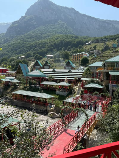 The image shows a resort area nestled in a valley with a mountain range in the background. The resort features several multi-story buildings, many with green, sloped roofs, and covered seating areas. A river flows through the foreground, crossed by a bright red bridge with glass panels and decorative floral arches. Several people are visible walking on the bridge and in the seating areas, some of whom appear to be tourists. The vegetation is lush and green, indicating a natural, possibly mountainous, environment. The overall impression is of a scenic and recreational destination. The location is identified as Qırızdəhnə, Azerbaijan.
