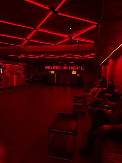 The image shows an indoor space illuminated with red light, likely a club or bar. Neon lights are arranged in geometric patterns on one wall, while the words "MUSIC IS HOPE" are vertically displayed in red neon on another. People are seated in a row of chairs along the wall, some with tables in front of them. The seating appears to be a live music venue in Guadalajara, Mexico.