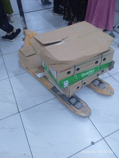 The image shows a pallet jack with several cardboard boxes stacked on it. The boxes are of various sizes and appear to be made of brown cardboard. Some of the boxes have green labels with white text, including "Qualiko" and "Cherkizovo." The pallet jack itself is yellow and appears to be made of metal. It has wheels, and the forks are extended to lift the boxes. The floor is tiled with white and grey tiles. In the background, people are visible, wearing various clothing items. One person is wearing a long green skirt, another dark pants and black shoes, and another a pink dress. A portion of a shopping cart is also visible on the right side of the image. The scene is likely indoors, possibly in a storage or retail environment. The timestamp "2026/6/29 12:07" is visible in the bottom right corner. There is no specific indication of the location being Ak-Suu, Kyrgyzstan.