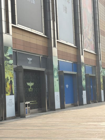 The image captures a vertical view of a building facade in Yuhuatai District, Nanjing, Jiangsu, China. The building features a series of commercial units, each identifiable by large signs. The top unit displays images of food, likely advertising a restaurant. The unit below contains a large blank, likely an advertisement. Below that is a unit prominently displaying the logo of Adidas, a multinational corporation that designs and manufactures athletic shoes and clothing. The next unit features the brand "Boy London," indicating a fashion retail outlet. Beneath it is another large advertisement. The architecture is characterized by a modular design, with each unit framed by metal structures, and a neutral color palette of creams, browns, and grays. The angle of the shot suggests a street-level perspective, with the building extending upwards. The sky is not visible, focusing attention on the urban environment and the building's commercial presence.