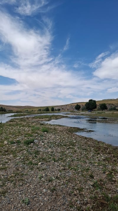The image depicts a wide, shallow river with numerous gravel bars and patches of green vegetation. The river flows through a gently rolling, arid landscape with sparse trees scattered along the banks. In the distance, hills rise, covered in dry grass and some rock formations. The sky is a vibrant blue with wispy white clouds. A blue car is visible parked on the right bank, partially obscured by trees. There are no people clearly visible in the image. The scene appears to be a natural, undeveloped area. Given the context of Boraldai, Kazakhstan, this is likely a view of a river or stream in the surrounding natural landscape.